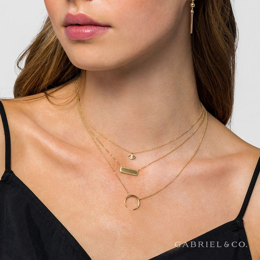A woman exudes personalized elegance in layered gold necklaces, featuring Gabriel & Co’s 14K Yellow Gold Rectangular ID Pendant Necklace with a twisted rope frame, paired with a gold earring and black spaghetti strap top.