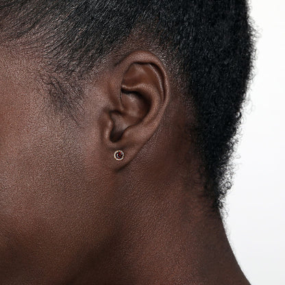 Close-up of an ear with short, curly hair wearing Gabriel & Co 14K Yellow Gold Round Halo Garnet and Diamond Stud Earrings, featuring a dark garnet center stone encircled by a sparkling diamond halo against a plain light background.