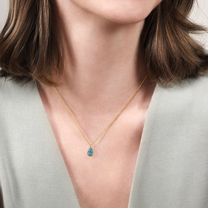 A woman with shoulder-length brown hair wears a light gray V-neck top and the Gabriel & Co 14K Yellow Gold Teardrop Blue Topaz Pendant Necklace—an elegant December birthstone gift.