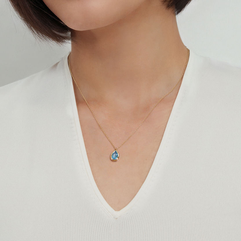 A woman wearing a white V-neck top and a Gabriel & Co 14K Yellow Gold Teardrop Blue Topaz Pendant Necklace—an ideal December birthstone gift. Photo is cropped to show only her neck and upper chest.
