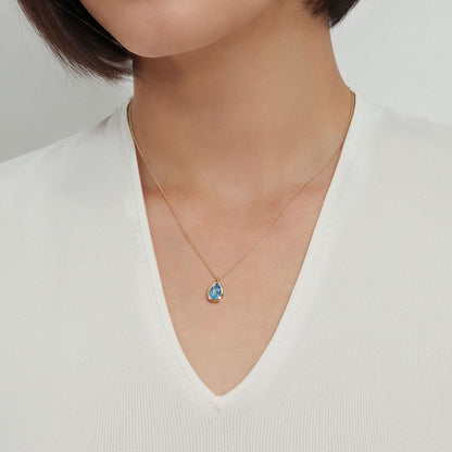 A woman wearing a white V-neck top and a Gabriel & Co 14K Yellow Gold Teardrop Blue Topaz Pendant Necklace—an ideal December birthstone gift. Photo is cropped to show only her neck and upper chest.