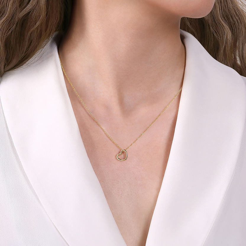 A woman in a white blazer showcases the Gabriel & Co 14K Yellow Gold Tilted Heart Pendant Necklace, featuring a delicate tilted heart resting at the center of her chest.