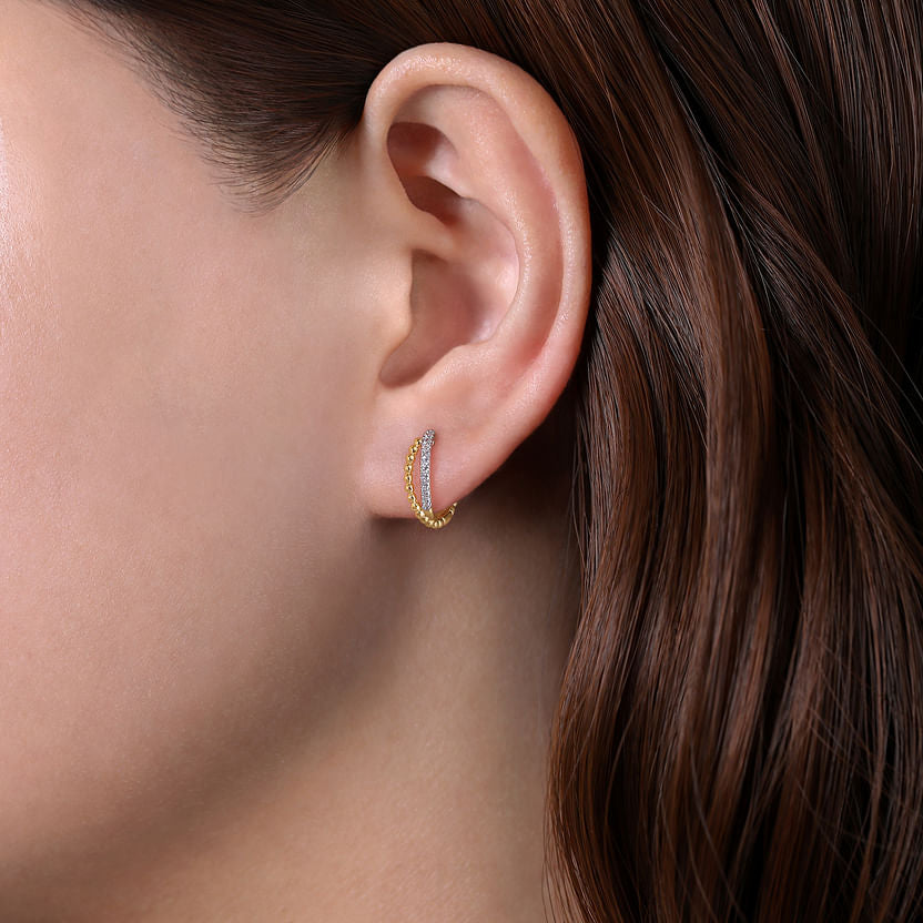 Close-up of a person’s ear wearing Gabriel & Co 14K Yellow Gold Twisted Pave 10mm Diamond Huggie Earrings, with straight brown hair tucked behind the ear.