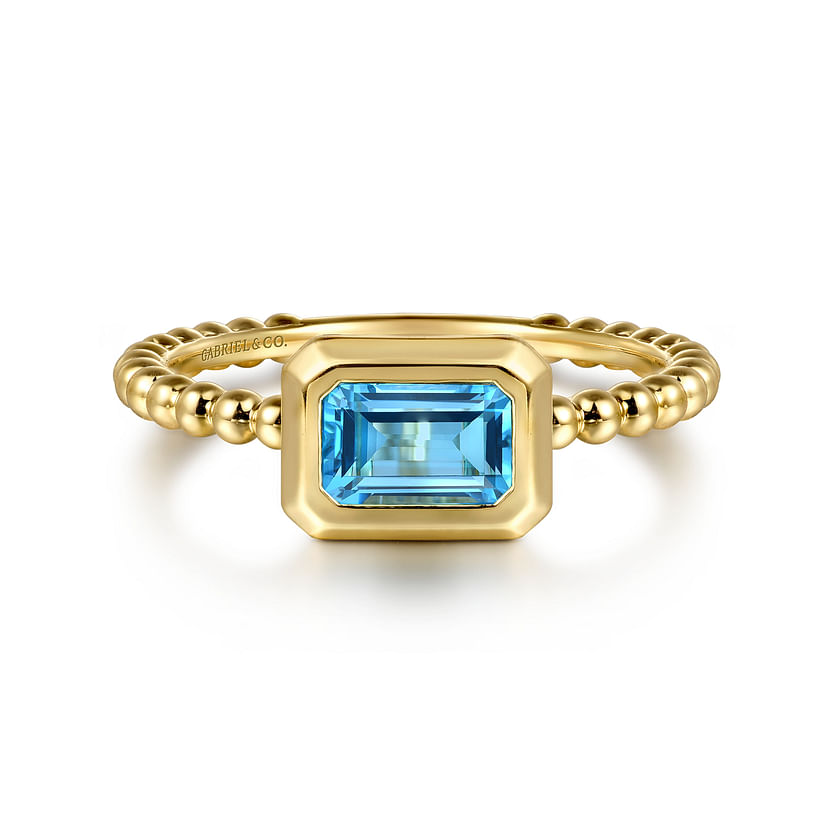 A 14K yellow gold Bujukan ring by Gabriel & Co, featuring a beaded band and rectangular blue topaz in a bezel setting, displayed on a white background. Stackable and stylish.