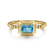 A 14K yellow gold Bujukan ring by Gabriel & Co, featuring a beaded band and rectangular blue topaz in a bezel setting, displayed on a white background. Stackable and stylish.