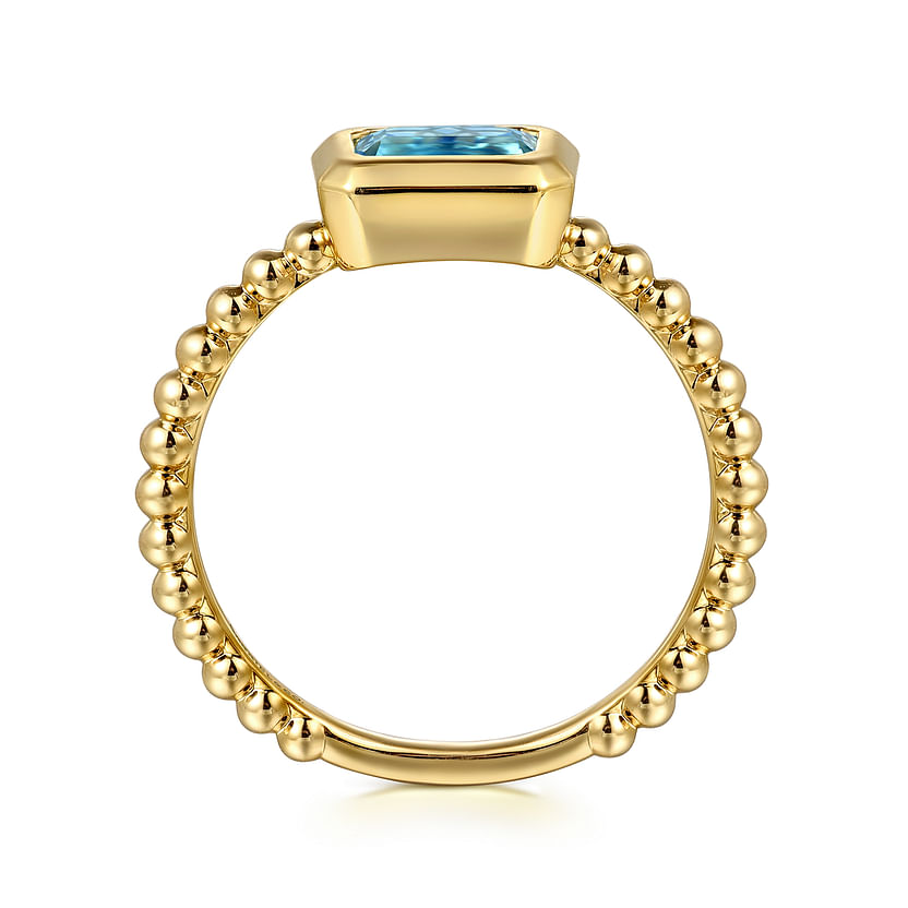 The Gabriel & Co 14K Yellow Plain Gold Bujukan and Blue Topaz Stackable Ring features a beaded gold band and a horizontal rectangular blue topaz, perfect for stacking, available at Avonlea Jewelers.