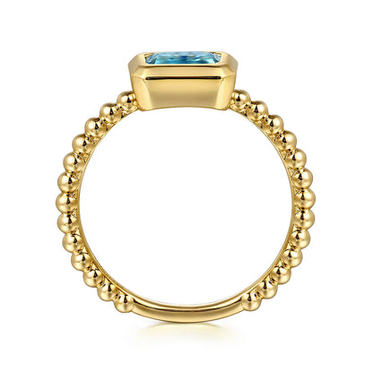 The Gabriel & Co 14K Yellow Plain Gold Bujukan and Blue Topaz Stackable Ring features a beaded gold band and a horizontal rectangular blue topaz, perfect for stacking, available at Avonlea Jewelers.