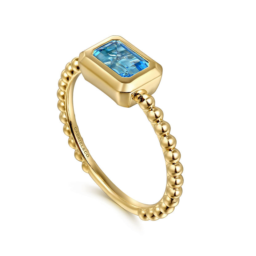 Discover the Gabriel & Co 14K Yellow Plain Gold Bujukan and Blue Topaz Stackable Ring—a gold beaded band with a rectangular blue topaz bezel set stone—available exclusively at Avonlea Jewelers.