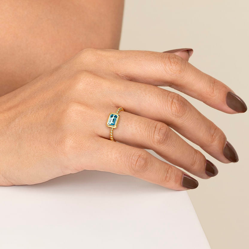 A hand with brown painted nails wears the Gabriel & Co 14K Yellow Plain Gold Bujukan and Blue Topaz Stackable Ring, featuring a rectangular blue gemstone, resting gently on a white surface.