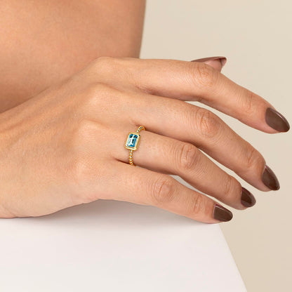 A hand with brown painted nails wears the Gabriel & Co 14K Yellow Plain Gold Bujukan and Blue Topaz Stackable Ring, featuring a rectangular blue gemstone, resting gently on a white surface.