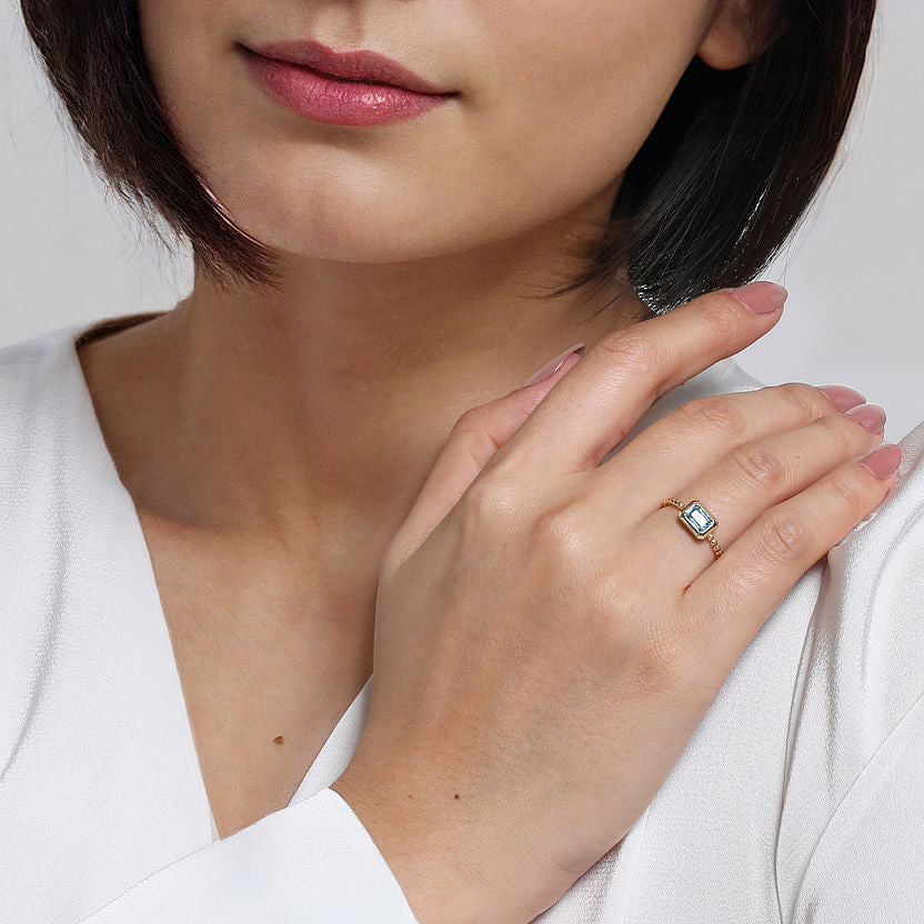 A woman with short dark hair in a white top gently rests her hand on her shoulder, showcasing the 14K Yellow Plain Gold Bujukan and Blue Topaz Stackable Ring by Gabriel & Co. Her neatly manicured nails are painted light pink.