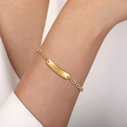 A close-up of a wrist adorned with the Gabriel & Co 14K Yellow Plain Gold Hollow Paperclip Chain Personalized ID Bracelet, featuring a sleek rectangular nameplate and thin chain. The wearer has on a light-colored long sleeve shirt.