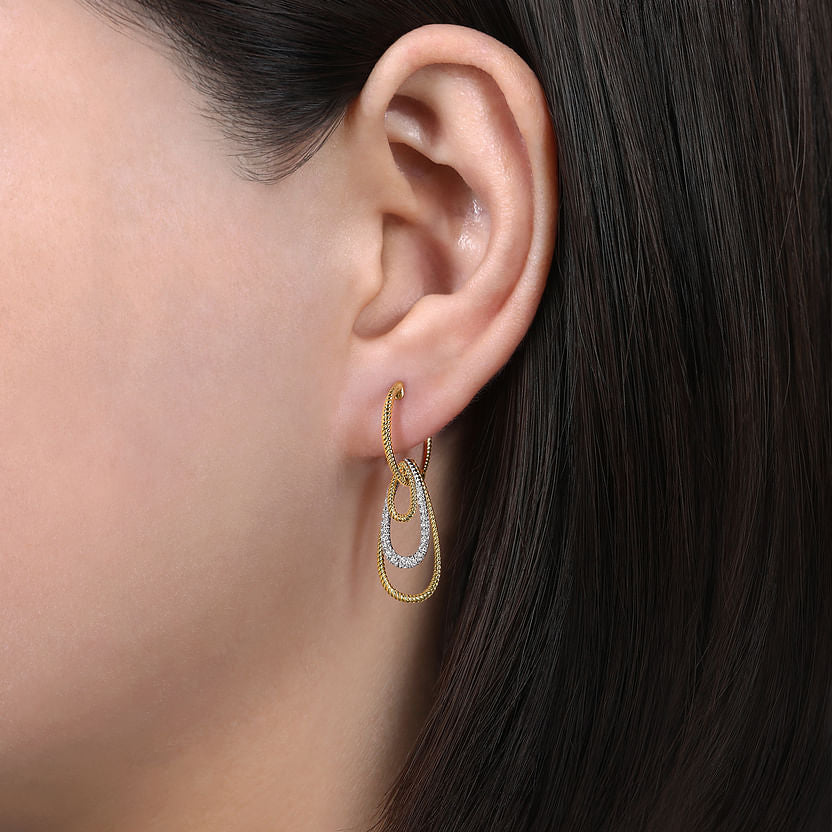 Close-up of a woman's ear with straight, dark brown hair, wearing Gabriel & Co 14K Yellow-White Gold Diamond Huggie Earrings featuring an elegant graduating teardrops design.