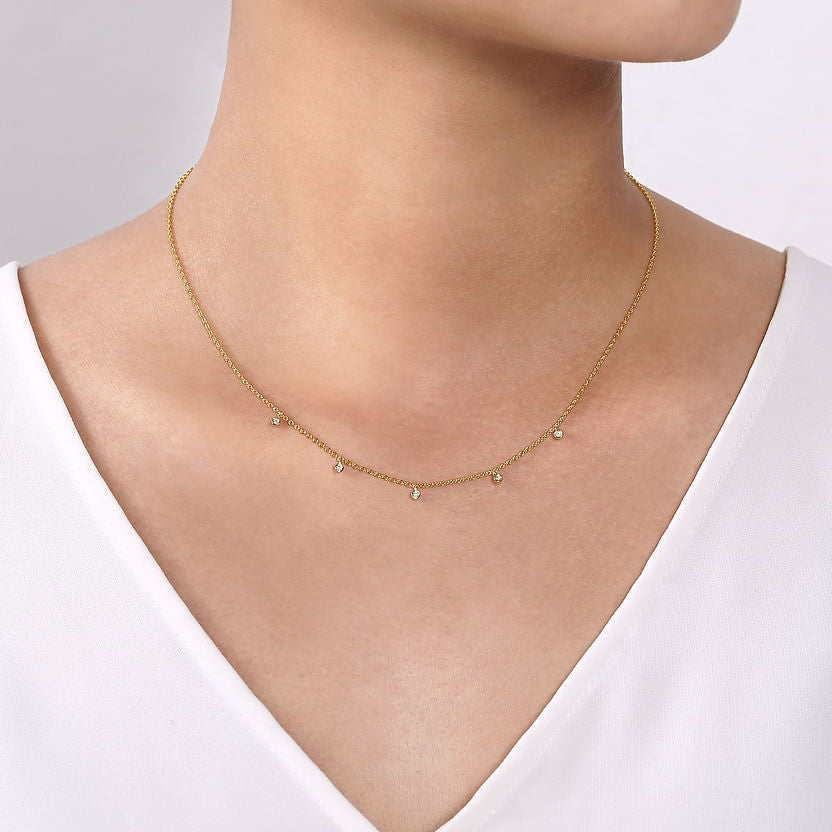 A close-up of a person in a white V-neck top wearing the Gabriel & Co 14K Yellow-White Gold Diamond Stations Droplet Necklace, a contemporary piece shown against a plain, neutral background.