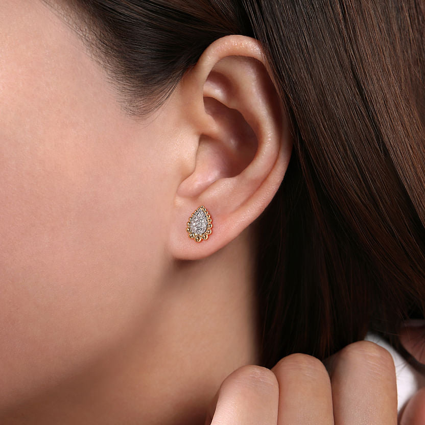 Close-up of a woman's ear with her brown hair tucked back, showcasing Gabriel & Co's 14K Yellow-White Gold Teardrop Shape Beaded Frame Diamond Stud Earrings and their elegant design.