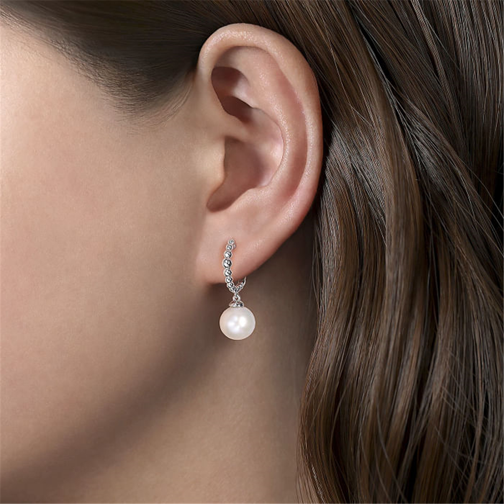 Close-up of a woman's ear adorned with Gabriel & Co's 925 Sterling Silver Bujukan Pearls Drop Huggie Earrings from the Bujukan Collection. The earrings feature a series of small, sparkling stones leading to a single, large Bujukan Pearl. Her hair is brown and straight, partially covering the ear. The background is blurred and neutral.