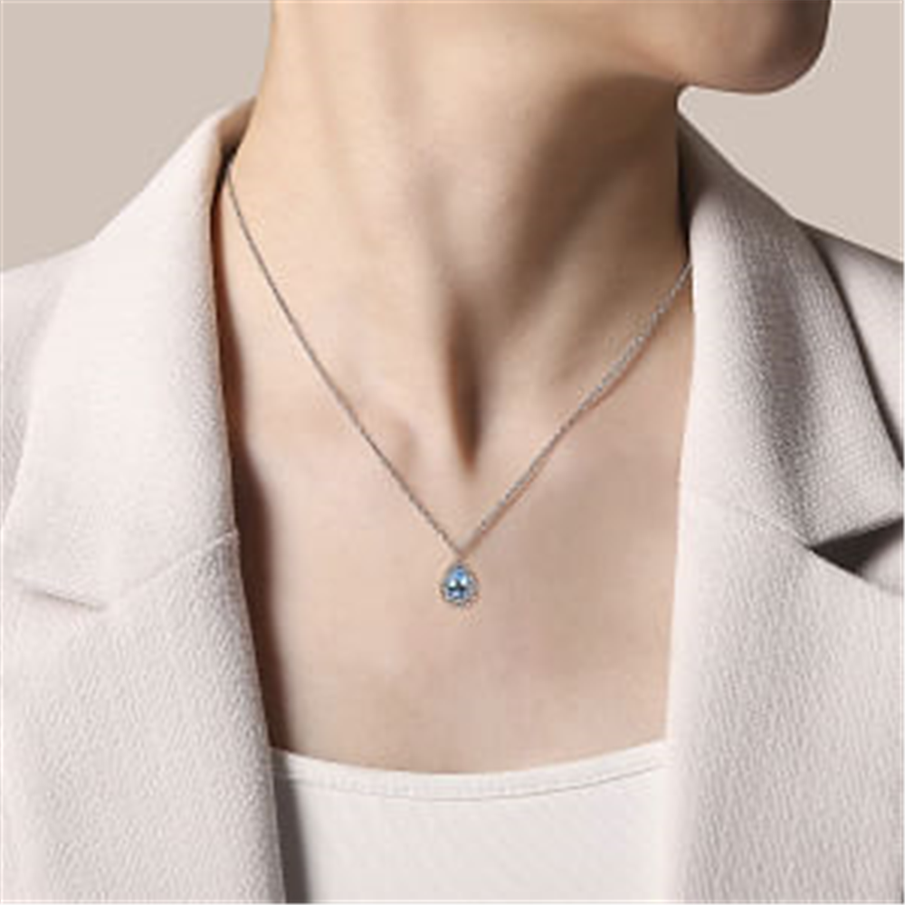 Close-up of a person wearing a white blazer and a Gabriel & Co 925 Sterling Silver Faceted Swiss Blue Topaz Pear Shape Bujukan Neck pendant on a delicate chain necklace. The person's head is turned slightly to the side, elegantly showcasing the beautiful pendant against the white fabric of the blazer. The background is plain and neutral.