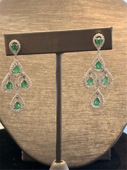 The Ryan Gems Diamond and Pear Shaped Emerald Dangles in 14k white gold feature teardrop green gemstones accented by sparkling stones, reminiscent of diamond earrings, displayed on a brown stand against a neutral textured background.