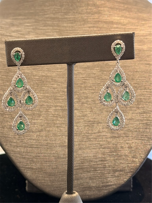 The Ryan Gems Diamond and Pear Shaped Emerald Dangles in 14k white gold feature teardrop green gemstones accented by sparkling stones, reminiscent of diamond earrings, displayed on a brown stand against a neutral textured background.