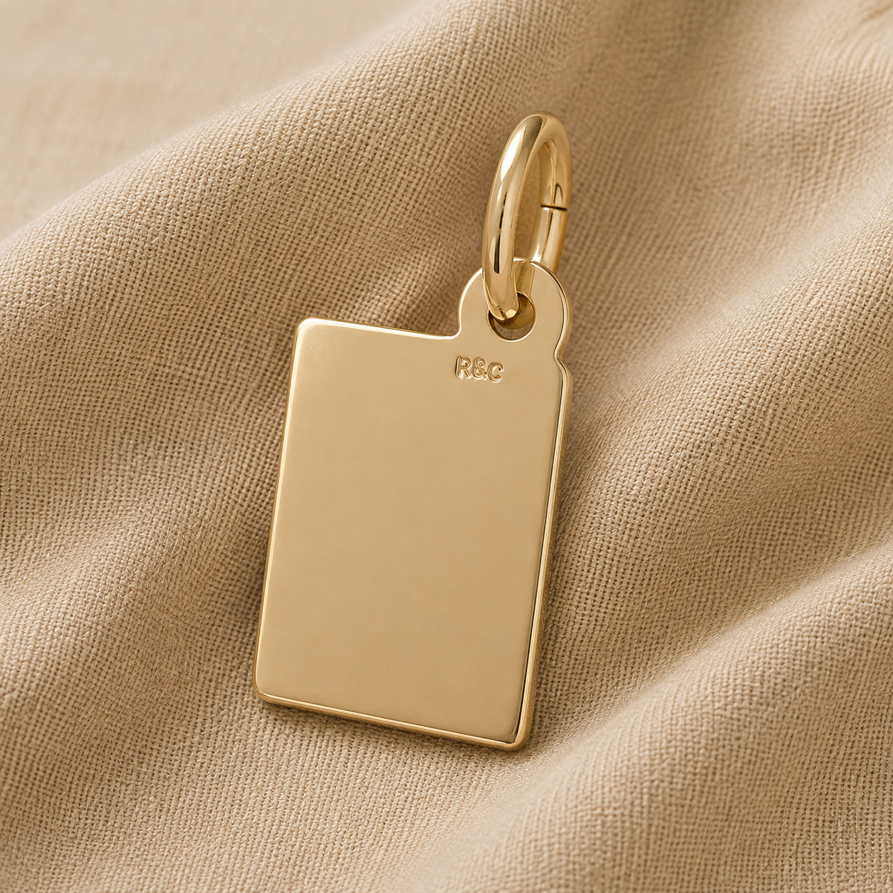 A gold-plated rectangular Book Charm from Rembrandt Charms, engraved with the initials “R&C” near the top, rests on soft beige fabric. This elegant Engravable Book Charm is a beautiful literary keepsake—perfect for anyone who cherishes personalized book jewelry.