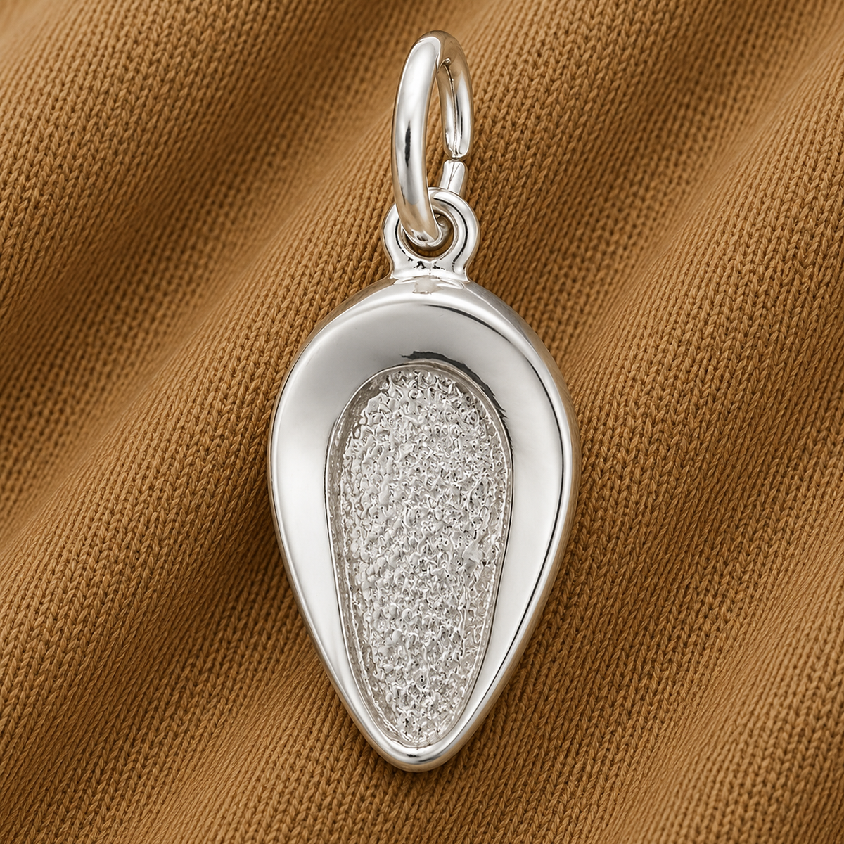 A gleaming sterling silver Bed Pan Charm by Rembrandt Charms, featuring an organic, oval shape with smooth edges and a textured center, rests atop a brown textured fabric background. With its large top loop for easy attachment to a chain, this unique piece makes an ideal healthcare jewelry accessory or thoughtful nurse gift.
