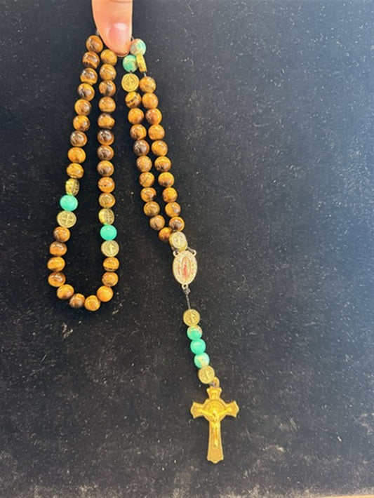 A hand holds the Redemption Jewelry LR002 Long Tiger Eye & Jade Rosary, featuring brown tiger eye and turquoise jade beads, a small medallion, and a gold crucifix at the end, set against a black background.