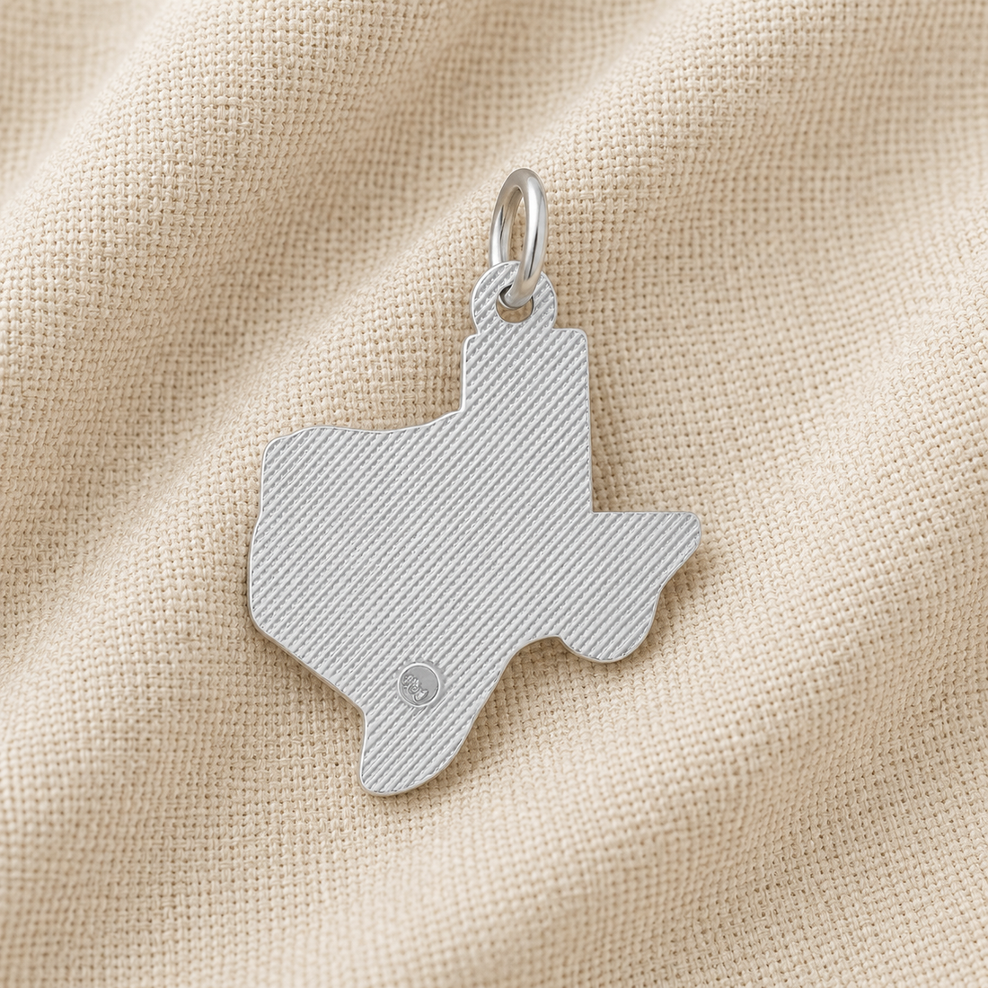 A Rembrandt Charms Texas State Map Charm in sterling silver rests on beige textured fabric. This distinctive Lone Star State pendant features fine diagonal lines and a small circular detail near the bottom, making it a unique piece of Texas jewelry.