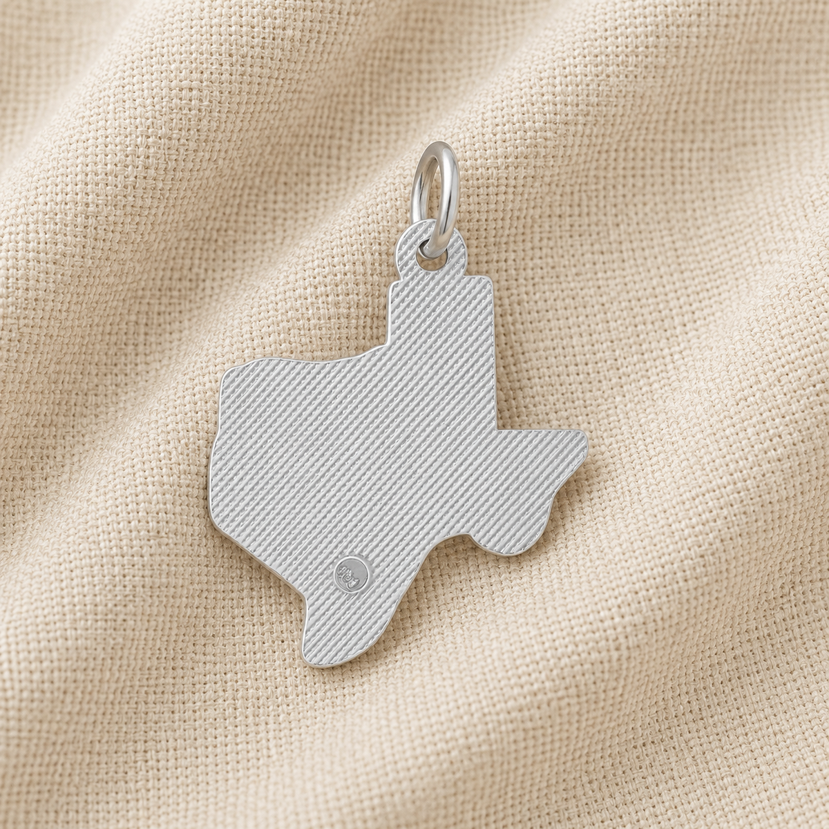 A Rembrandt Charms Texas State Map Charm in sterling silver rests on beige textured fabric. This distinctive Lone Star State pendant features fine diagonal lines and a small circular detail near the bottom, making it a unique piece of Texas jewelry.