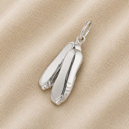 A Ballet Shoes -Sterling Silver charm by Rembrandt Charms, ideal as ballet jewelry or a cherished dance keepsake, rests on a textured beige fabric background.