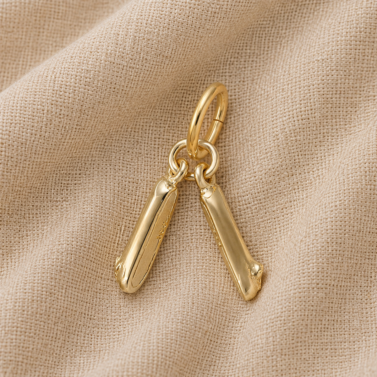 A gold-plated Pair of Ballet Shoes Charm by Rembrandt Charms, featuring two elongated, rounded charms reminiscent of ballet slippers, hangs from a small loop and rests on beige textured fabric.