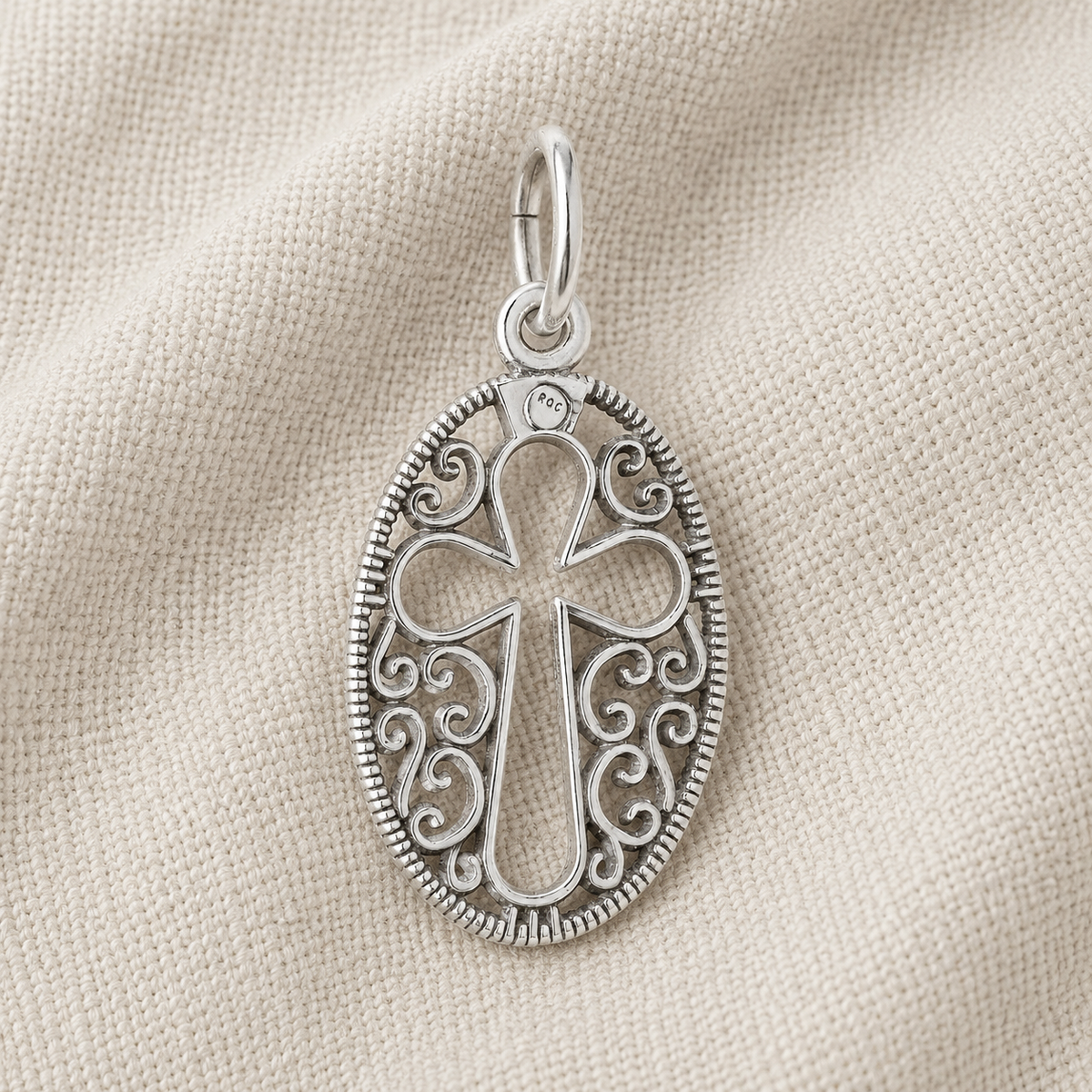 A gold-plated Filigree Oval Cross Charm by Rembrandt Charms features an openwork cross design with decorative scroll detailing, displayed on a textured beige fabric background.