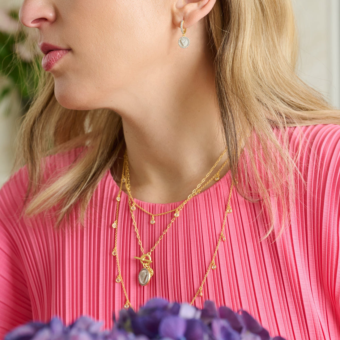 A woman in a bright pink pleated blouse wears a Sentiments Heart Charm Chain Link Necklace by Freida Rothman and heart-shaped earrings. Blurred purple flowers in the foreground enhance the romantic allure of her layered gold necklaces.