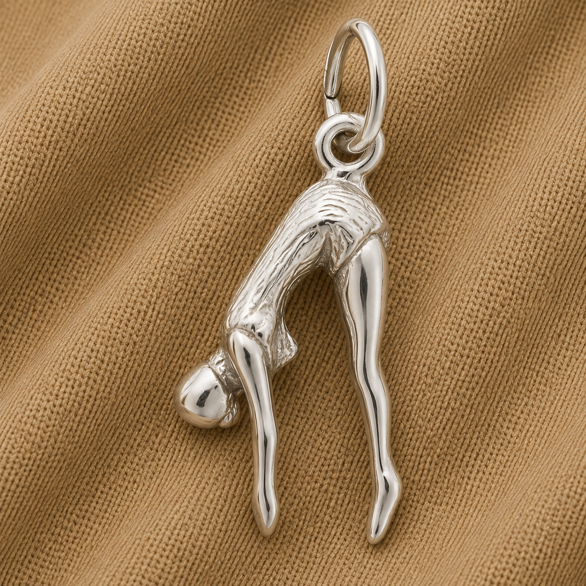 The Rembrandt Charms Diver Charm in sterling silver, shaped like a person bending forward and touching their toes, rests on light brown textured fabric—an ideal gift for swimmers who admire graceful movement and finely crafted details.