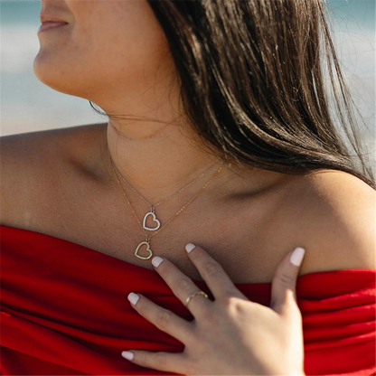 A woman with long dark hair wears a red off-the-shoulder top and the T Jazelle Beautiful Soul Gold Charm Necklace layered with a heart necklace. Her white-painted nails rest on her chest against a blurred beach background.