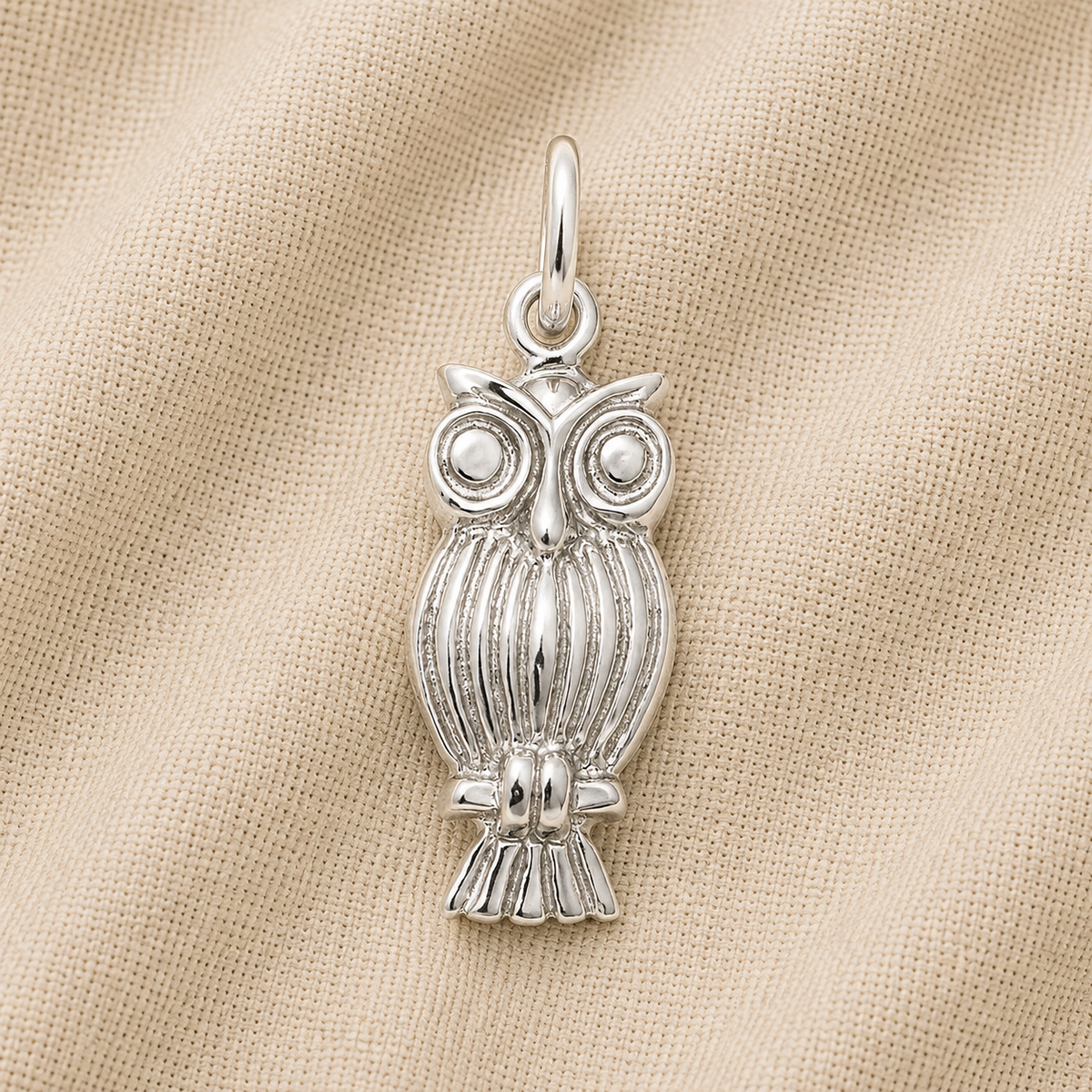 The Screech Owl Charm—Sterling Silver by Rembrandt Charms, featuring intricate feather detailing and expressive large eyes, rests on a soft beige textured fabric background, making it a perfect piece of owl jewelry and an ideal gift for nature lovers.