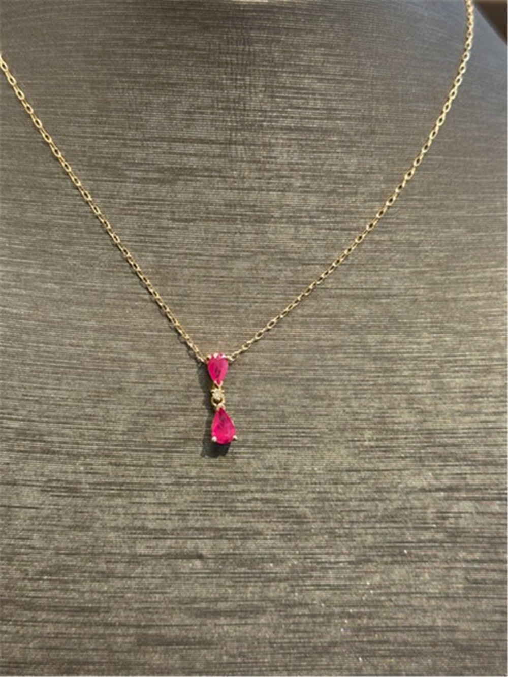 The Ryan Gems Ruby Drop Pendant features two pink rubies—an oval and a teardrop—set on a delicate 14k yellow gold chain, displayed on a gray textured jewelry bust.