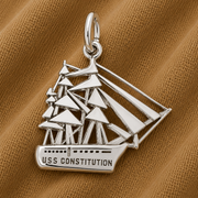 A sterling silver USS Constitution Charm by Rembrandt Charms rests on tan textured fabric with visible folds, showcasing its detailed sails and engraved ship name—a timeless piece of nautical jewelry that celebrates maritime heritage.