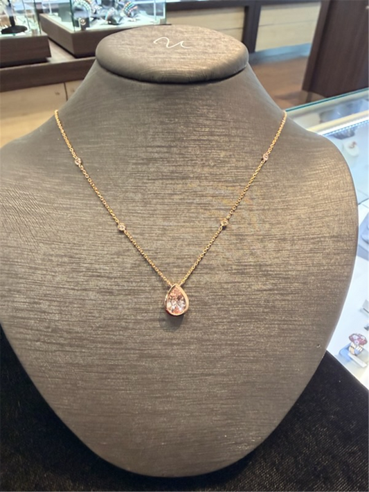 A Pear Shaped Morganite pendant by Ryan Gems, crafted in 14k rose gold and featuring a light pink gemstone, is displayed on a black jewelry bust in a store setting.
