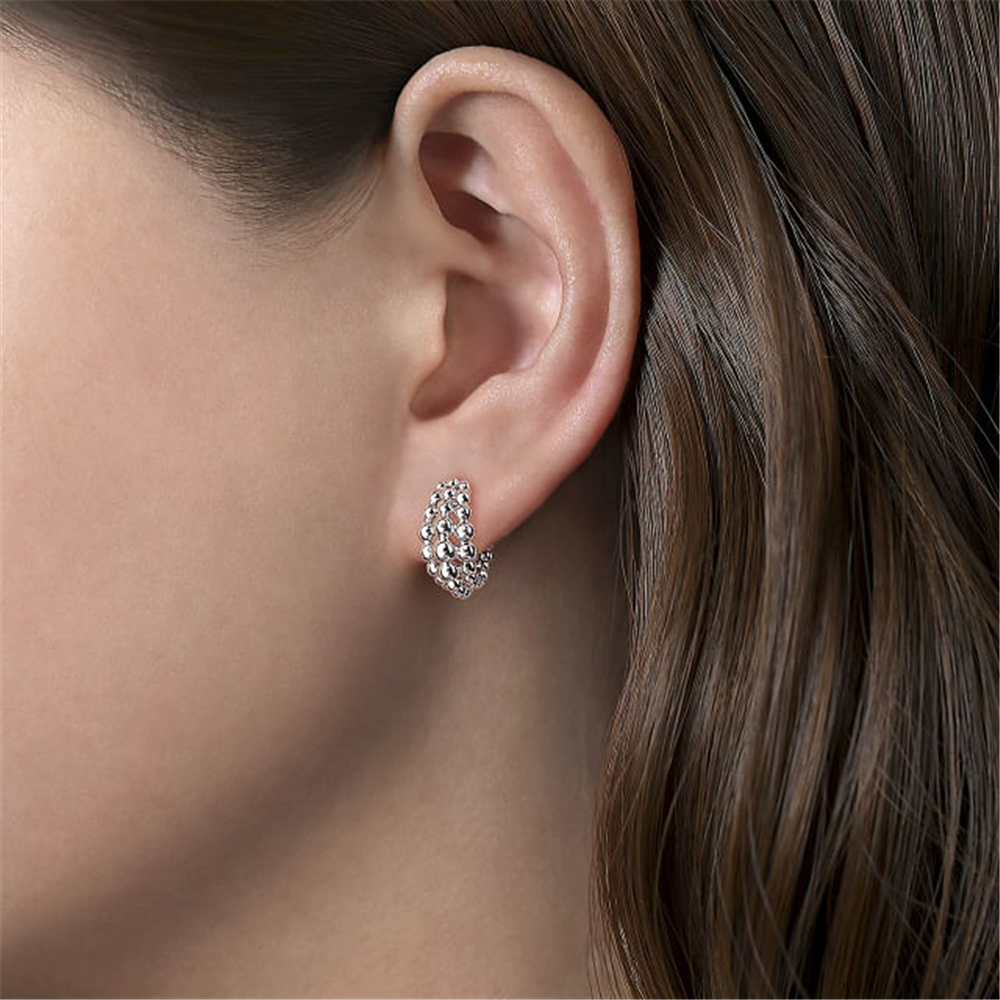 A close-up shot highlights a person's ear embellished with a white, beaded, textured earring from Gabriel & Co's Bujukan Collection. The individual has straight brown hair neatly tucked behind the ear, prominently displaying the 925 Sterling Silver Bujukan Huggie Earrings against a neutral backdrop.