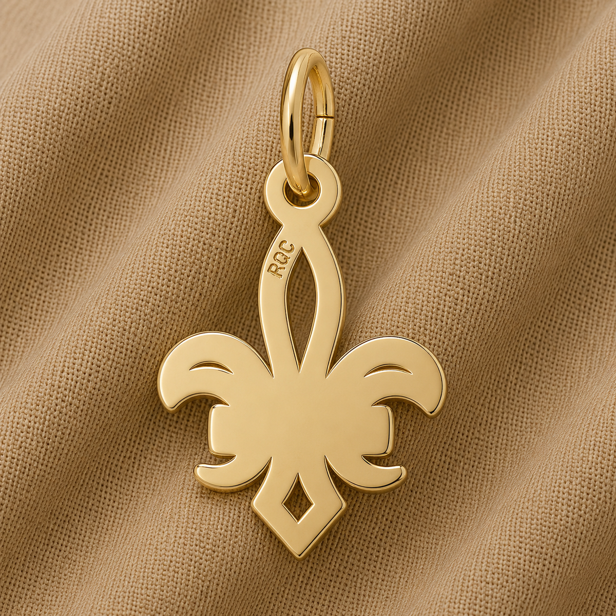 A gold-plated NOLA Fleur de Lis Charm by Rembrandt Charms, featuring a smooth, shiny surface and “RCC” engraved on one side, rests atop tan fabric—a timeless Crescent City keepsake and an elegant piece of New Orleans jewelry.
