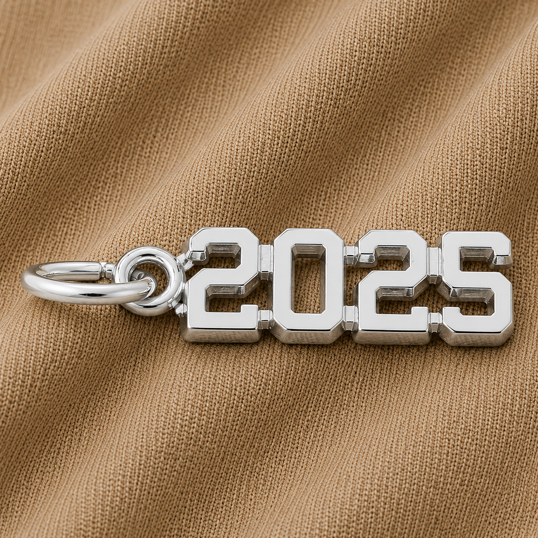 The Rembrandt Charms 2025 Graduation Charm—crafted in sterling silver—rests on a textured beige fabric background, making it an ideal graduation gift and a memorable keepsake for the class of 2025.