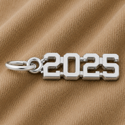 The Rembrandt Charms 2025 Graduation Charm—crafted in sterling silver—rests on a textured beige fabric background, making it an ideal graduation gift and a memorable keepsake for the class of 2025.