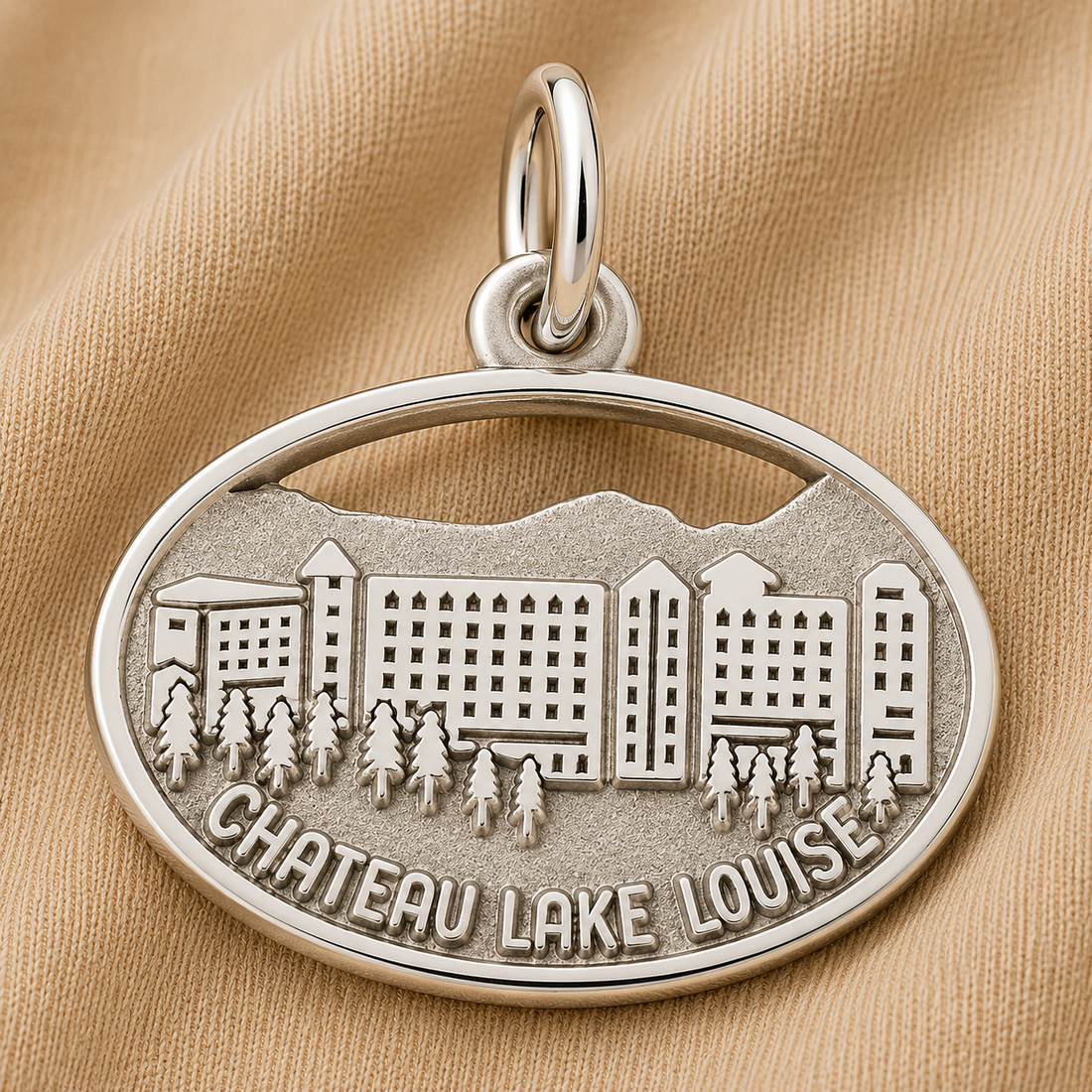 The Rembrandt Charms Chateau Lake Louise - Sterling Silver Charm, intricately engraved with buildings, trees, mountains, and the words “CHATEAU LAKE LOUISE,” beautifully captures the majesty of the Canadian Rockies as it rests on a beige fabric background.