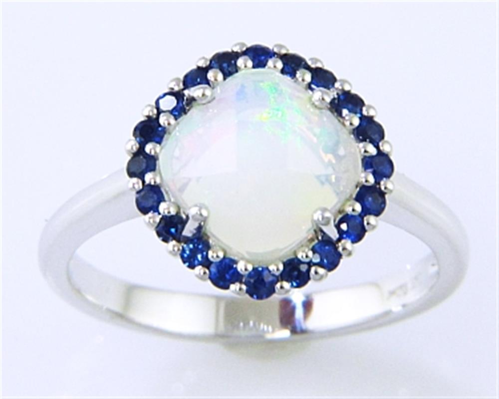 The Bellarri Faceted Opal and Blue Sapphire Ring features a 0.94ct round opal with an iridescent shine, encircled by small blue sapphires totaling 0.35ct, all set in polished 14KWG for a smooth and elegant finish.