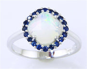 The Bellarri Faceted Opal and Blue Sapphire Ring features a 0.94ct round opal with an iridescent shine, encircled by small blue sapphires totaling 0.35ct, all set in polished 14KWG for a smooth and elegant finish.