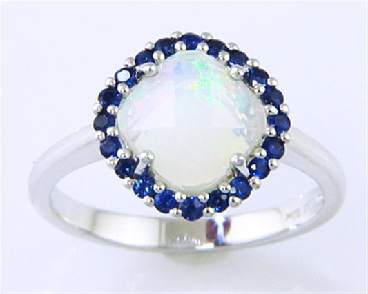 The Bellarri Faceted Opal and Blue Sapphire Ring features a 0.94ct round opal with an iridescent shine, encircled by small blue sapphires totaling 0.35ct, all set in polished 14KWG for a smooth and elegant finish.