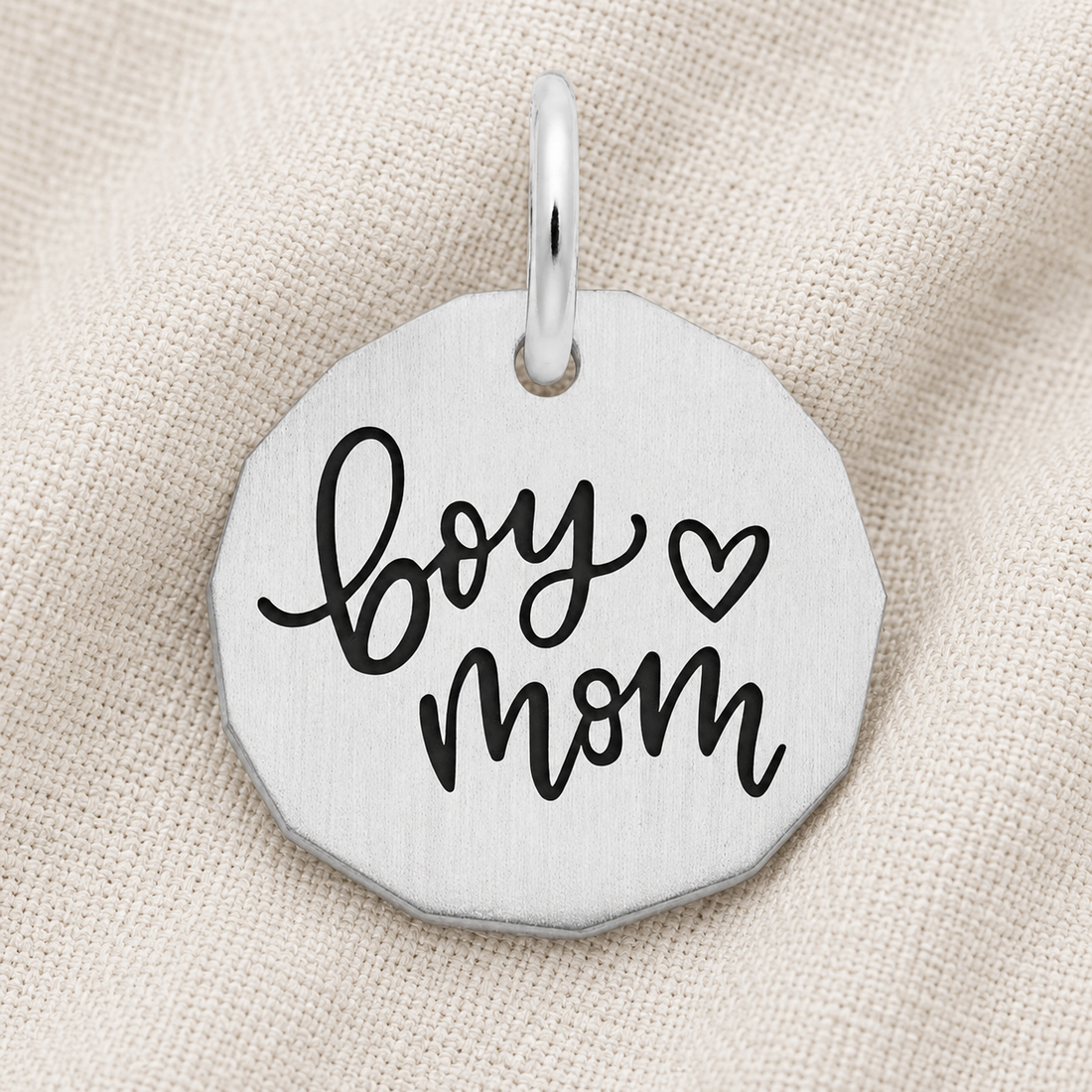 Introducing the Boy Mom Charm by Rembrandt Charms—a sterling silver pendant engraved with “boy mom” and a heart in bold black lettering, displayed on a beige textured fabric. This elegant charm is an ideal piece of mother and son jewelry or a thoughtful gift for Mother’s Day.