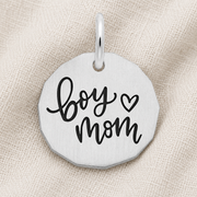 Introducing the Boy Mom Charm by Rembrandt Charms—a sterling silver pendant engraved with “boy mom” and a heart in bold black lettering, displayed on a beige textured fabric. This elegant charm is an ideal piece of mother and son jewelry or a thoughtful gift for Mother’s Day.