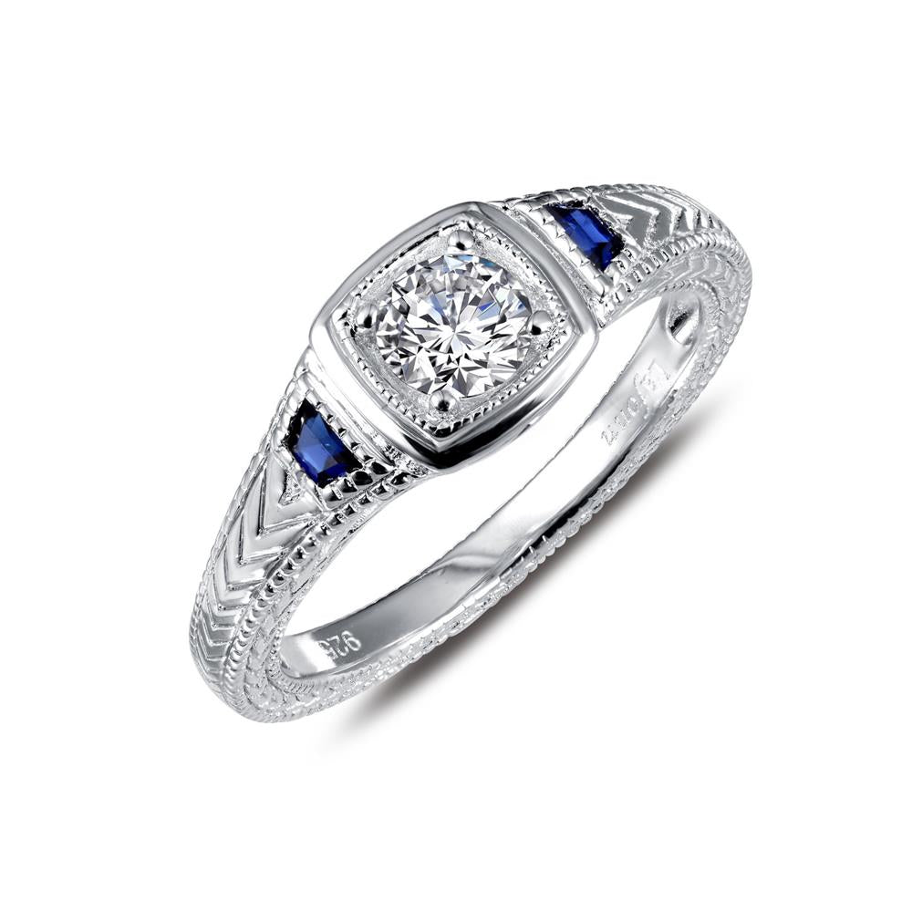 The Lafonn Art Deco Inspired Engagement Ring showcases a round central diamond set in a square frame, accented with blue gemstones on either side. The intricately decorated band is adorned with detailed engravings.