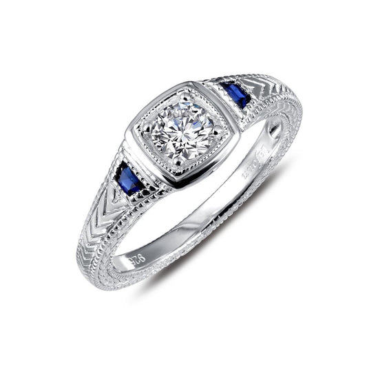 The Lafonn Art Deco Inspired Engagement Ring showcases a round central diamond set in a square frame, accented with blue gemstones on either side. The intricately decorated band is adorned with detailed engravings.